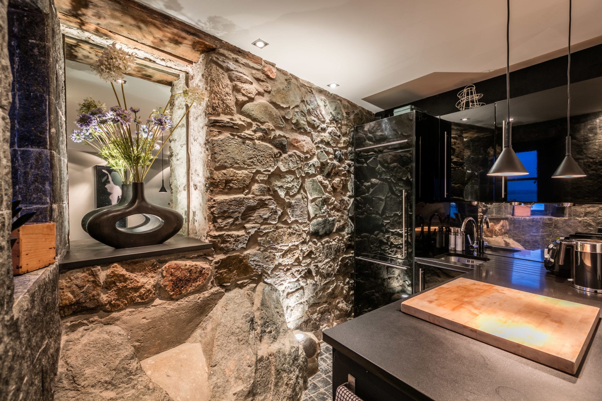 A kitchen with matte black surfaces, exposed stone and feature lighting