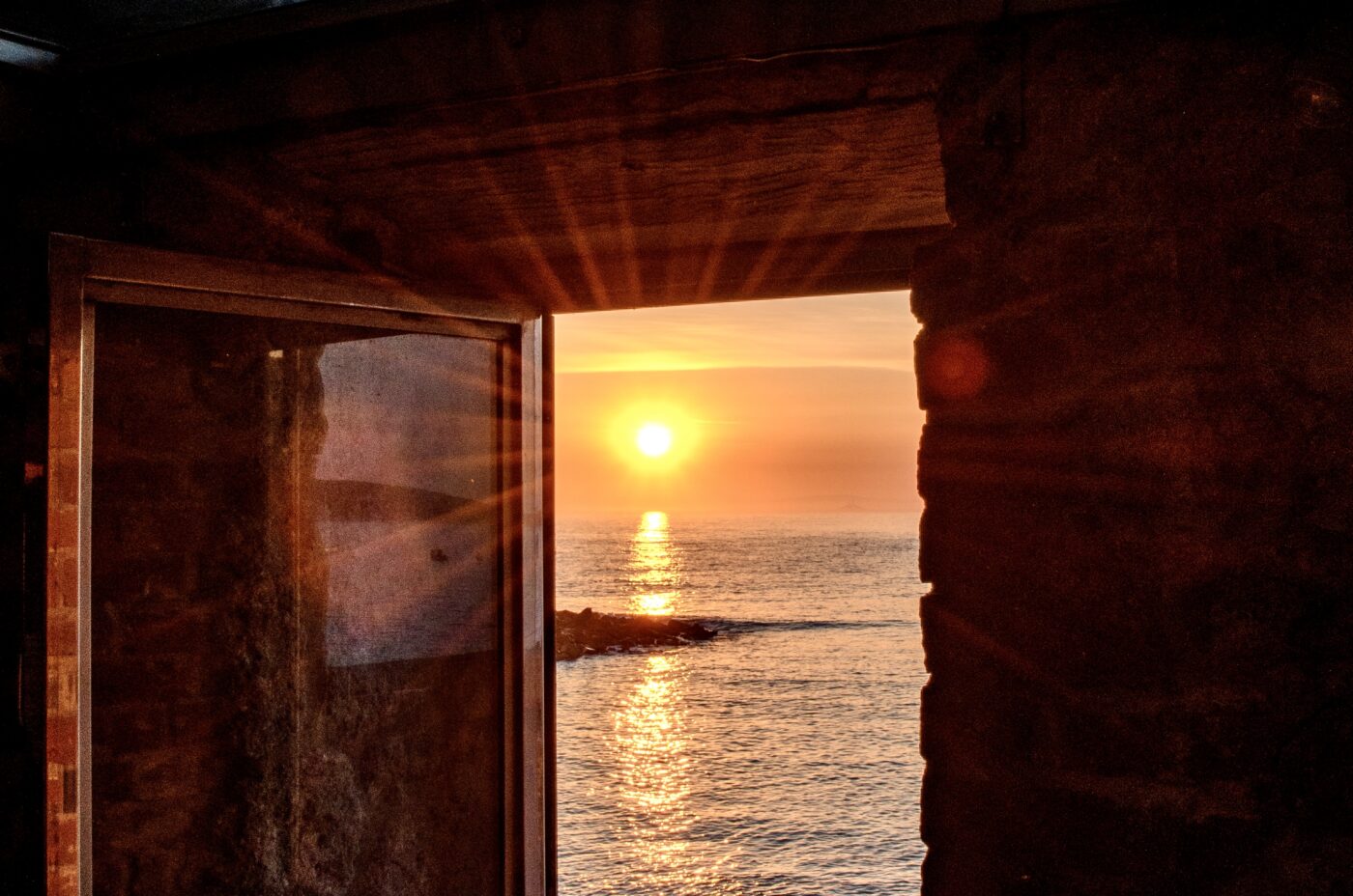 A small window in a stone wall with a view of the sunrise over the sea