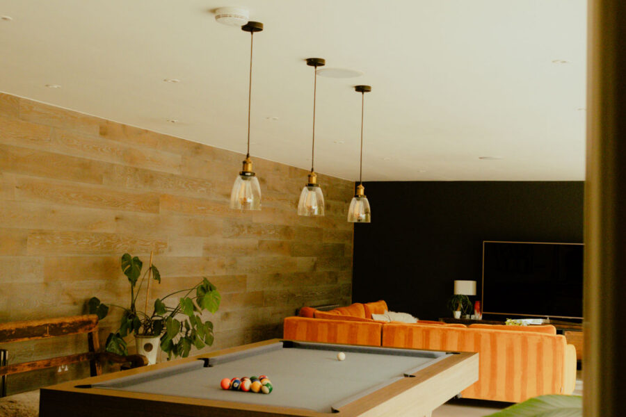 Games room with a pool table set up for a game, orange velvet sofas, a wood-paneled accent wall, and pendant lighting.