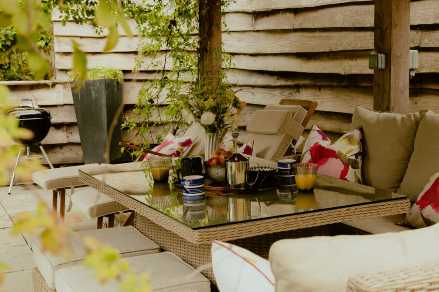 Wide view of the same covered patio, showing a barbecue, potted plant, wildflower bouquet, and glass table with breakfast items among wooden slatted walls.