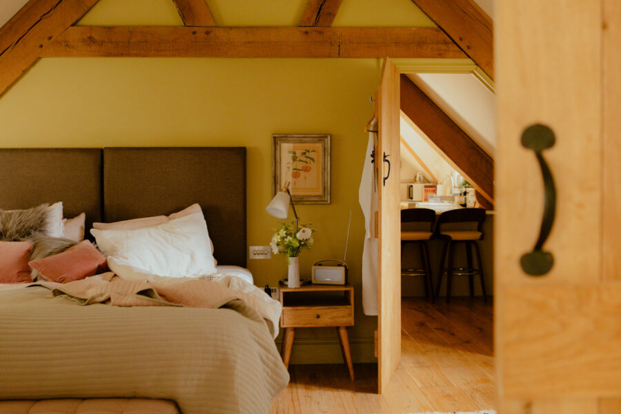 Bedroom with a plush bed and pink cushions under a sloped yellow ceiling with exposed beams, opening onto a kitchenette with bar stools.