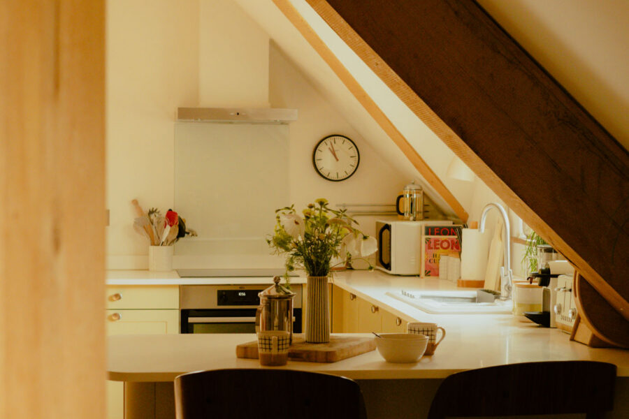 Attic kitchen with a breakfast bar and stools in the foreground, leading to a fitted kitchen with a wall clock, oven, and sink beneath a sloped beamed ceiling.