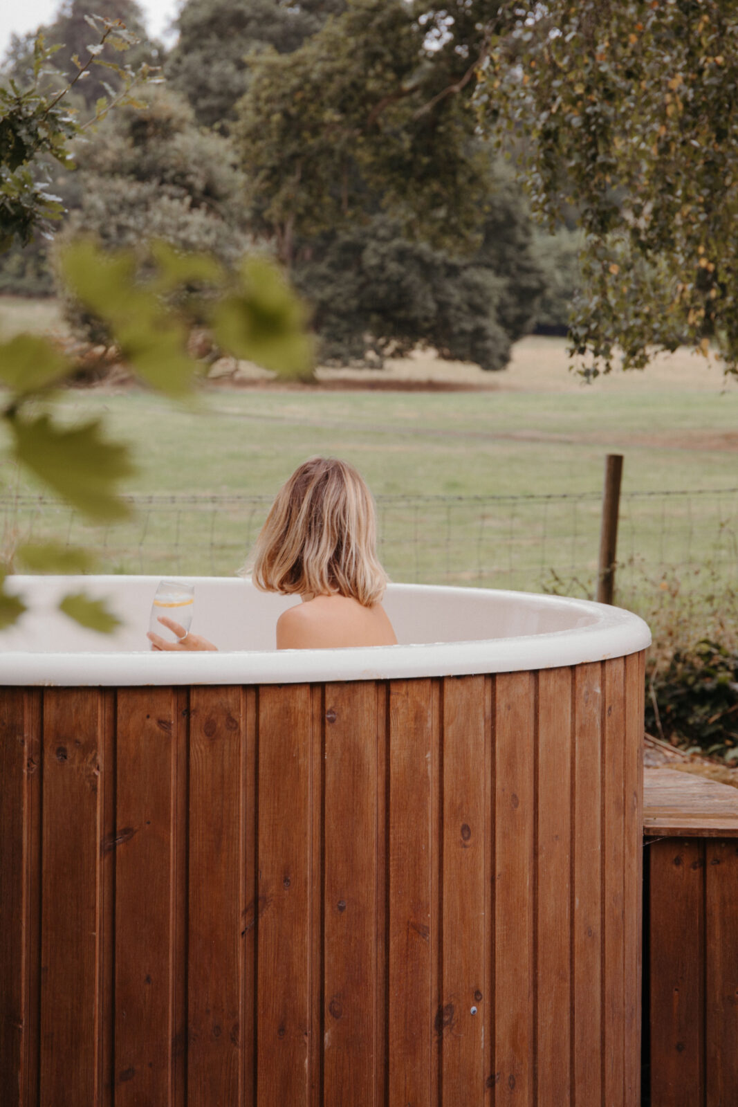 A woman with blonde hair in a wooden hot tub looks out to the nearby fields