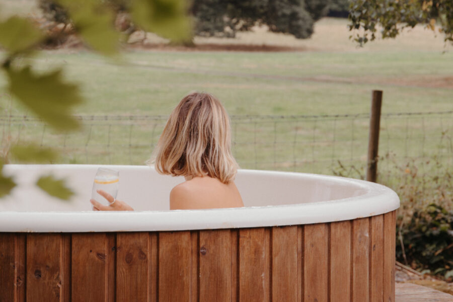 A woman with blonde hair in a wooden hot tub looks out to the nearby fields
