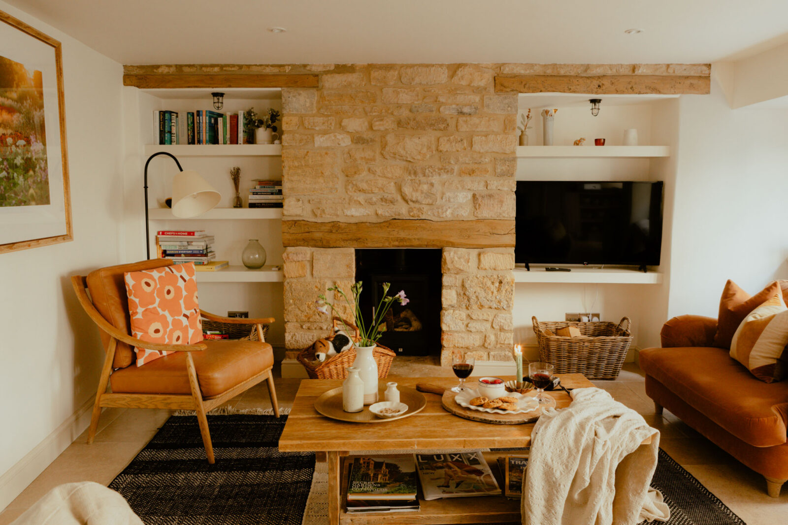 Cosy cottage sitting room with an exposed Cotswold stone fireplace and wood burner, a tan leather armchair and a rust velvet sofa.