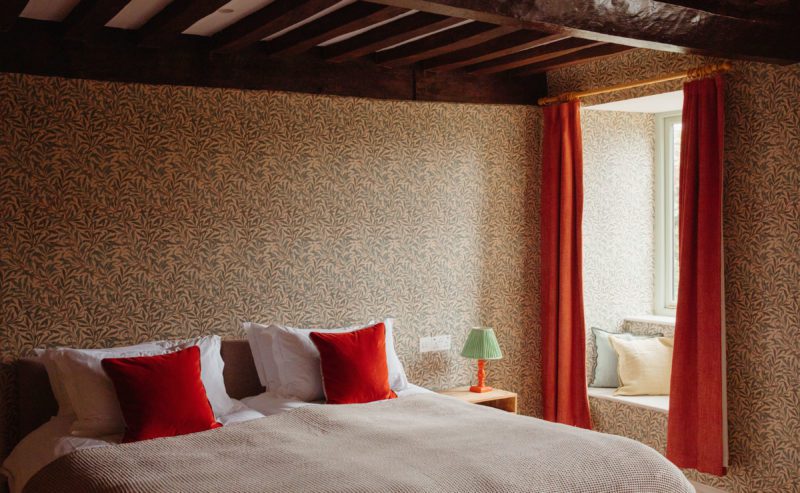 A bedroom with decorative wallpaper, wooden beams and red curtains