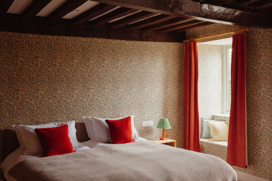A bedroom with decorative wallpaper, wooden beams and red curtains