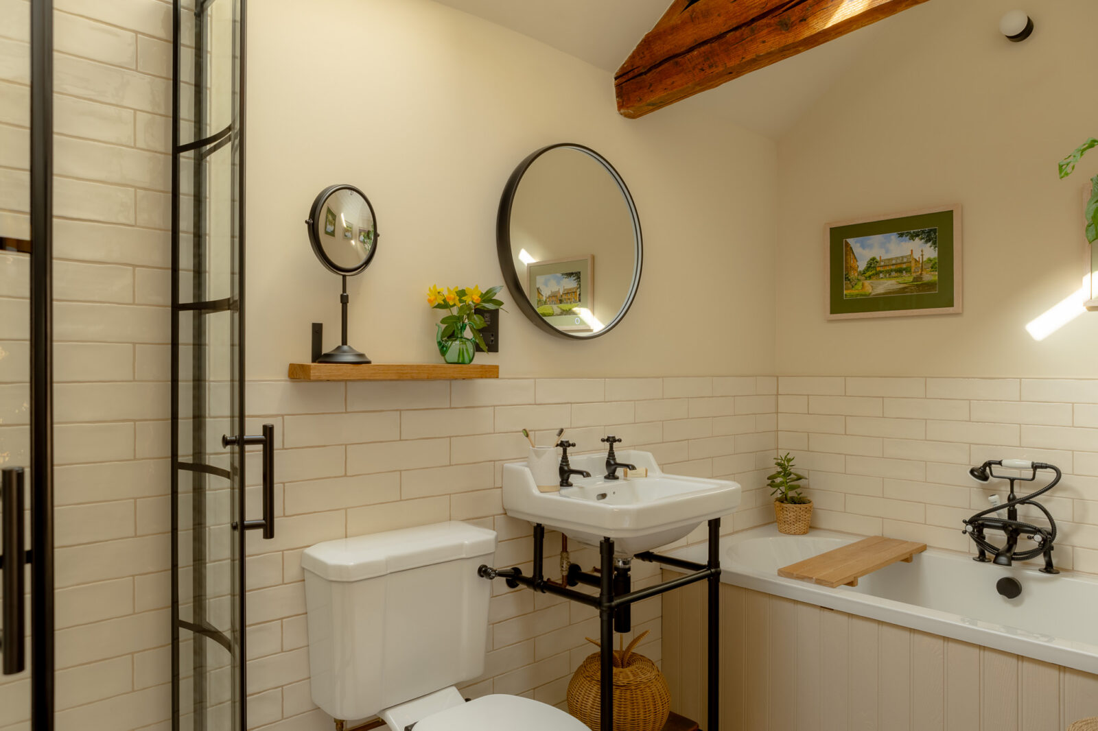 Bathroom showing a curved black-framed glass shower enclosure, white pedestal sink with black taps, close-coupled WC, cream subway tiles, round black-framed mirror, small oak shelf with vanity mirror and yellow flowers, and an exposed oak beam overhead.