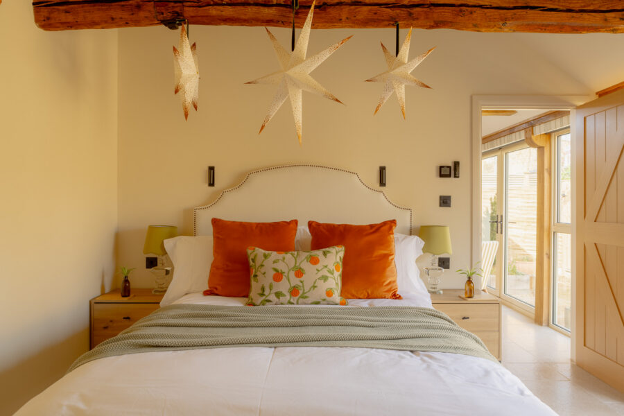 Bedroom with a cream upholstered king-size bed dressed in burnt-orange velvet cushions and a citrus-print accent cushion, sage waffle throw, flanking oak bedside chests with glass lamps, star-shaped pendant lights hanging from an exposed beam, and French doors opening to a private terrace.