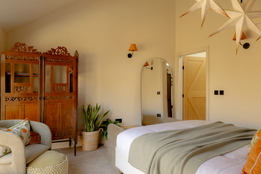 Spacious bedroom with high ceiling, a cream upholstered bed with sage waffle throw, an ornate carved wooden folding screen used as a wardrobe, a grey velvet armchair with citrus-print cushion, a full-length arched mirror, snake plant in a wicker basket, orange wall sconces, and a star-shaped pendant light.