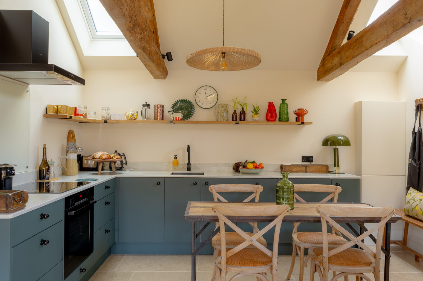 Open-plan kitchen with teal-green shaker cabinets, white marble-effect worktops, open oak shelving displaying colourful ceramics and vases, a rattan pendant light, exposed oak beams, Velux skylights, and a rustic wooden dining table with cross-back chairs.