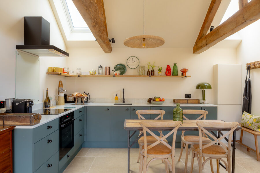 Open-plan kitchen with teal-green shaker cabinets, white marble-effect worktops, open oak shelving displaying colourful ceramics and vases, a rattan pendant light, exposed oak beams, Velux skylights, and a rustic wooden dining table with cross-back chairs.