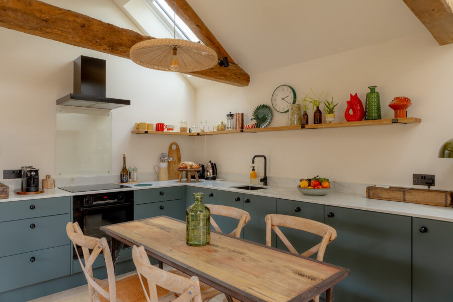 Open-plan kitchen-diner with teal shaker cabinets, marble-effect worktops, open oak shelving with colourful ceramics, a rattan conical pendant light, exposed A-frame oak beams, a Velux rooflight, and a reclaimed rustic dining table with cross-back chairs.