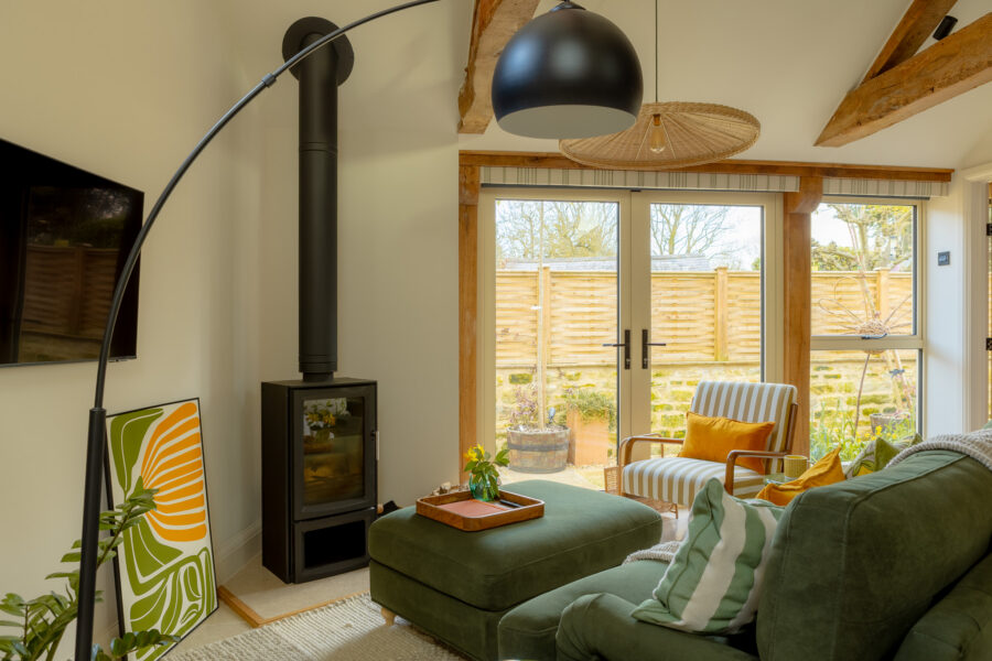 Living room featuring a black wood-burning stove with a dramatic tall flue, an oversized arc floor lamp, rattan pendant light, sage-green sofa and matching ottoman, French doors with garden views, a striped armchair on the terrace, and a retro green-and-orange botanical print.