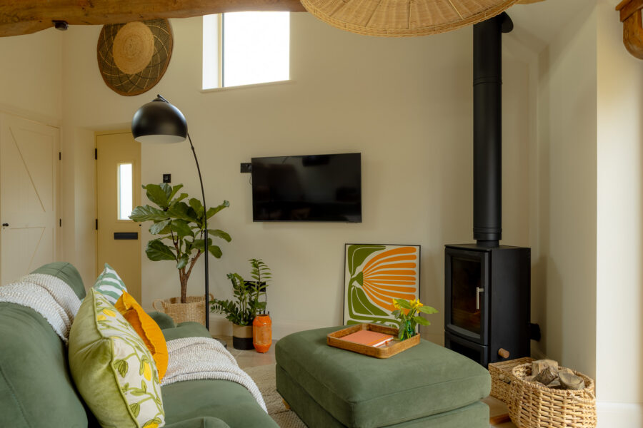 Living room with a sage-green velvet sofa and matching ottoman, black wood-burning stove with flue pipe, wall-mounted TV, large arc floor lamp, fiddle-leaf fig plant, rattan and wicker pendant light, exposed oak beam, and a bold green-and-orange abstract print.