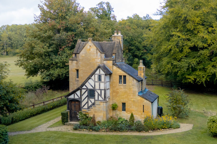 An aerial shot of a cotswold stone lodge with a Tudor style front in the middle of a secluded clearing with leafy trees and fields in the distance.