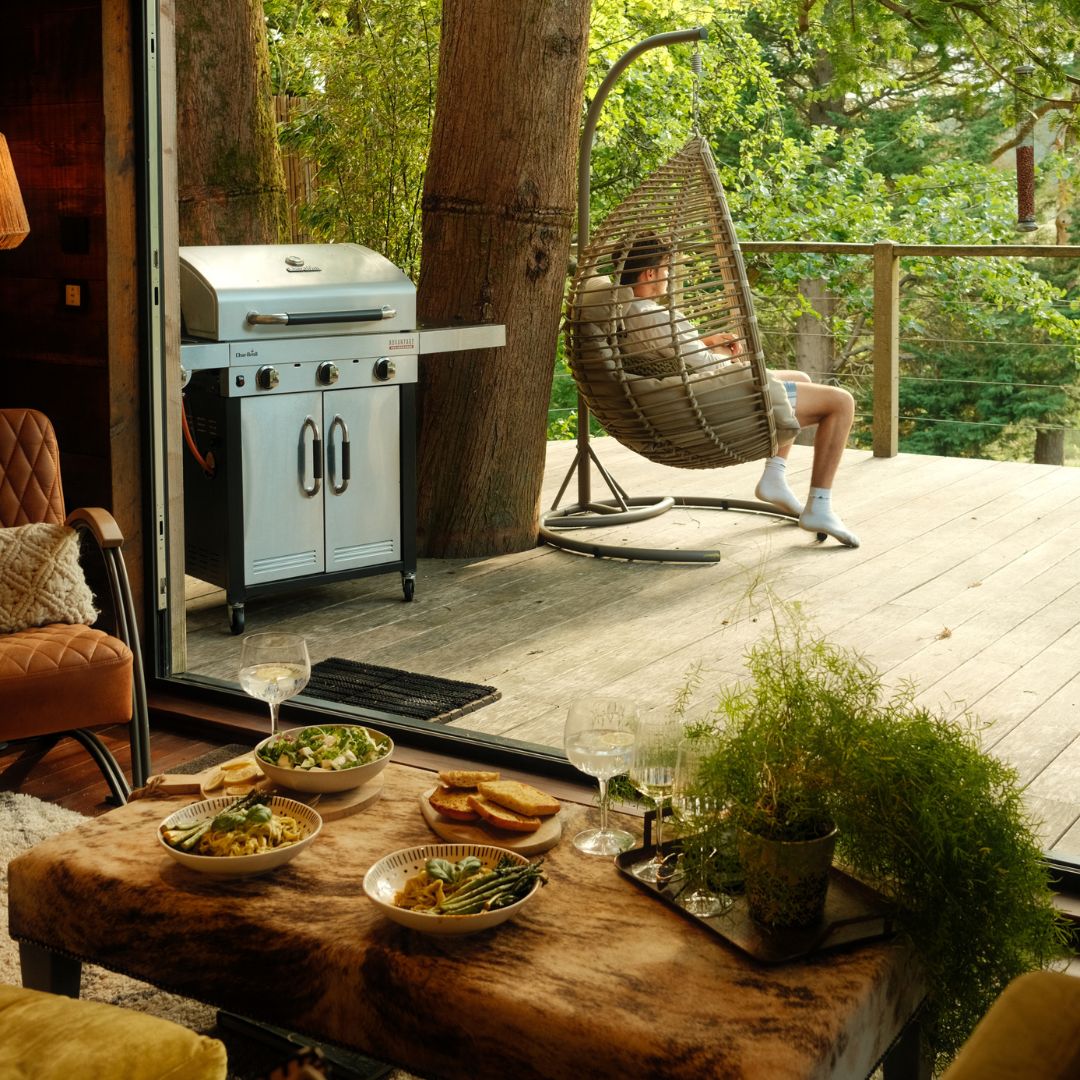 A man sits in a hammock chair on a deck with a BBQ and a table laden with food.
