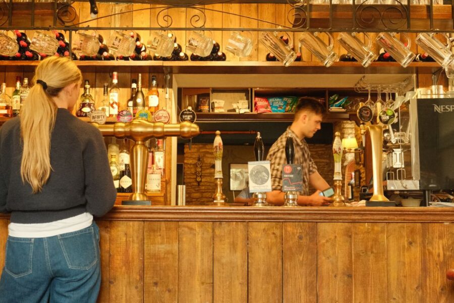 The back of a woman with blonde hair at the bar of a pub with the sign 'Cat & Custard Pot'