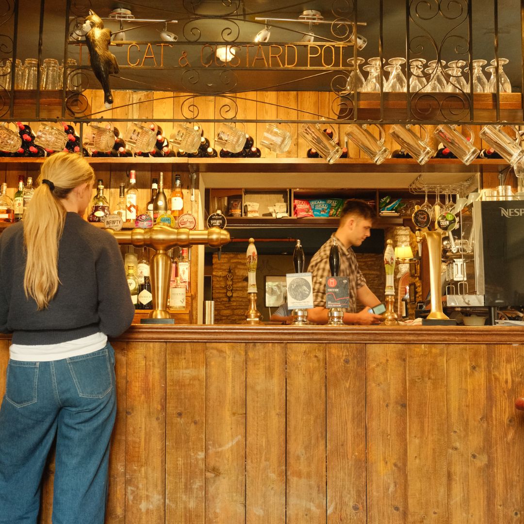 The back of a woman with blonde hair at the bar of a pub with the sign 'Cat & Custard Pot'