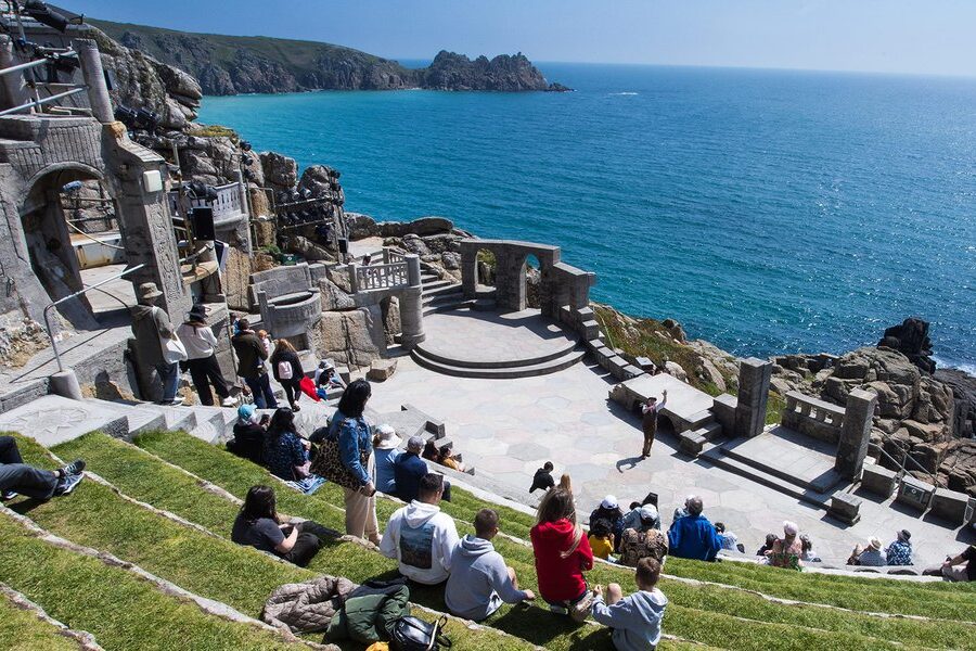 Open-air stone theatre built into the cliffside overlooking the sea with people beginning to gather.