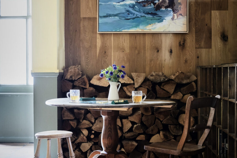 We wooden table, chair, and stool in a cosy corner with wooden panelled walls and a pretty seaside picture directly behind.