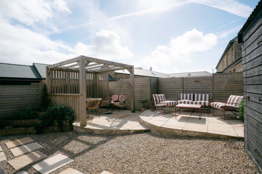 A outdoor seating set up underneath a wooden parasol with patio and gravel floor on a sunny day.