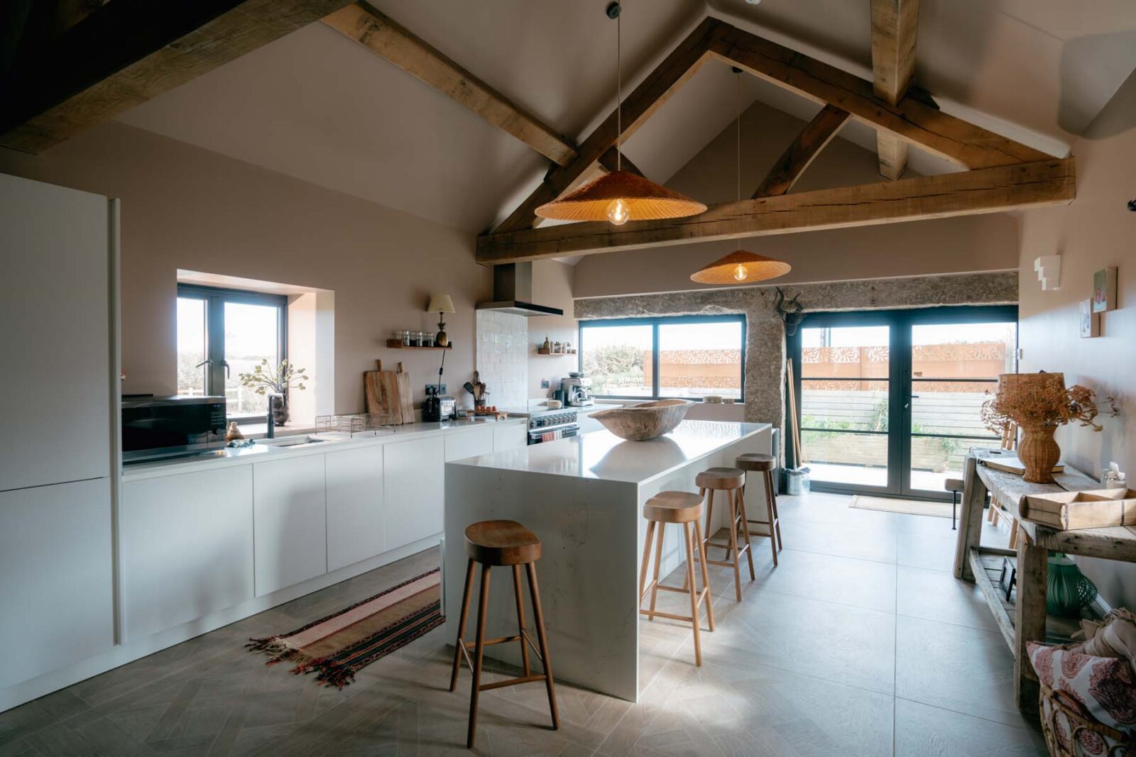 A modern kitchen with an island and stools with overhead beams and hanging pendant lights with floor-to-ceiling windows in the background.