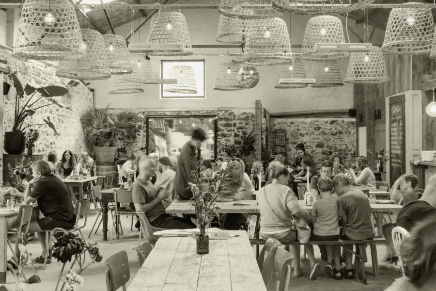 Busy restaurant set up with overhead hanging pendant lights and wooden long tables.
