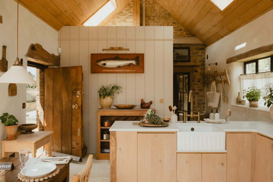 Vaulted kitchen-diner with exposed stone chimney breast, mounted fish artwork, oak island with butler sink and brass tap, and a reserved place setting in the foreground.