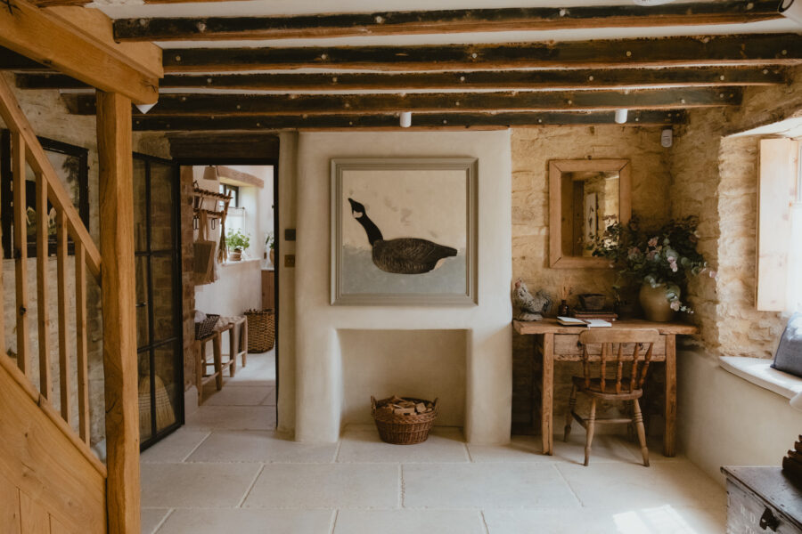 Ground floor hallway with oak staircase, a framed goose painting above the fireplace, a rustic writing desk with mirror, and a glimpse through to the kitchen beyond.