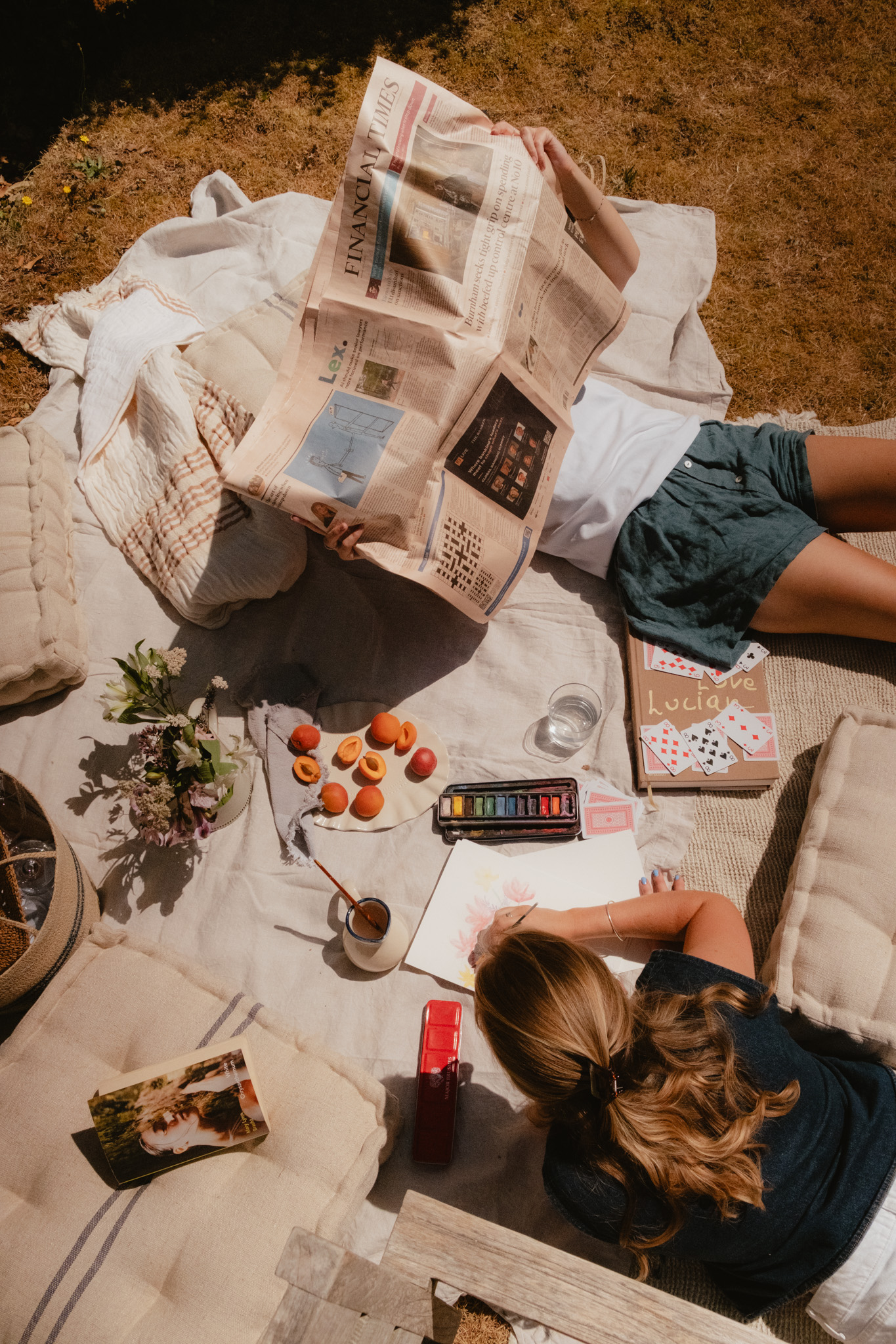 Overhead view of a woman reading the Financial Times on a picnic blanket beside watercolour painting supplies, playing cards, apricots and a magazine.