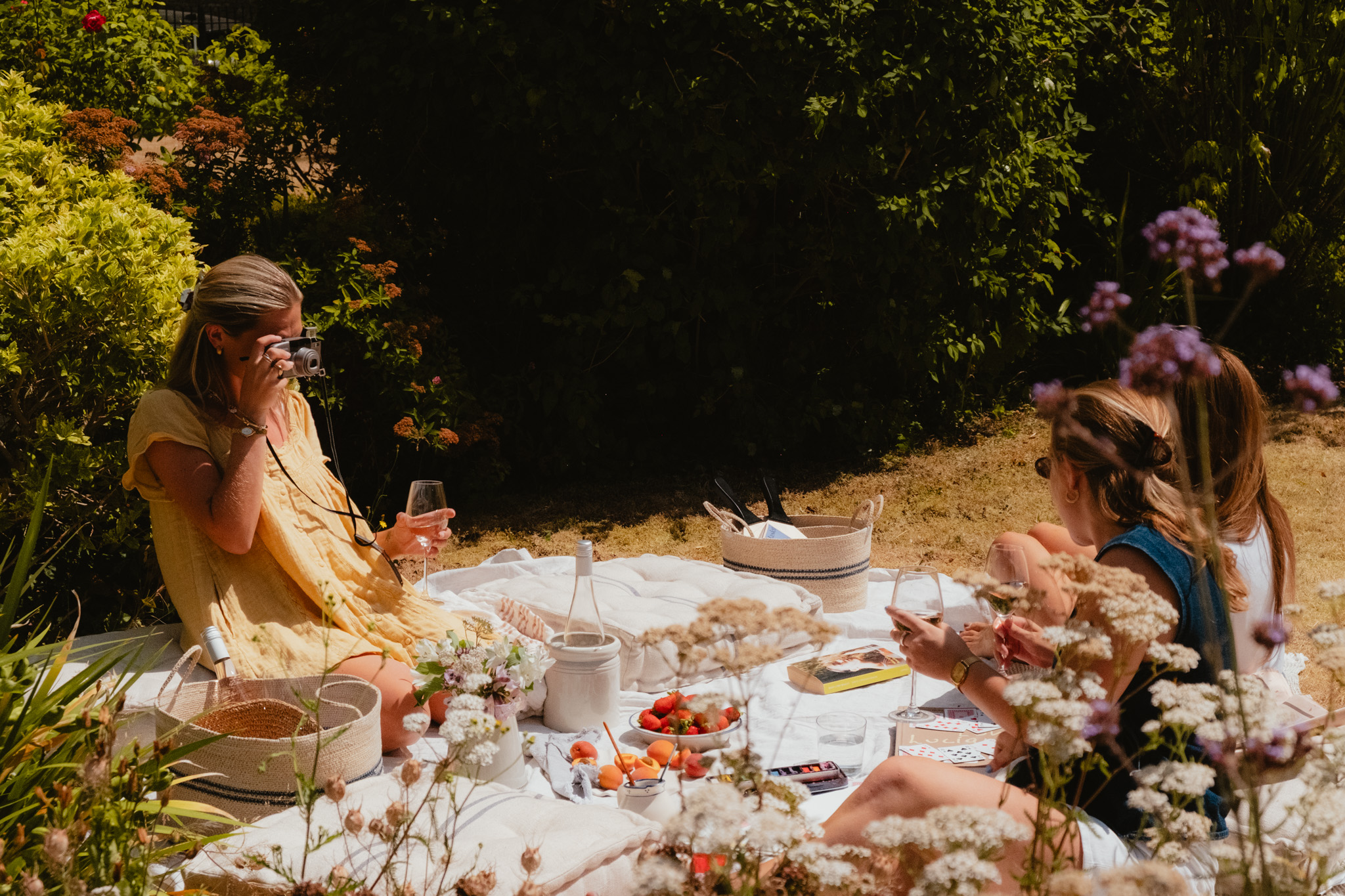 Three women relax on a picnic blanket in dappled shade, one taking a photograph with a compact camera while the others share wine and playing cards.