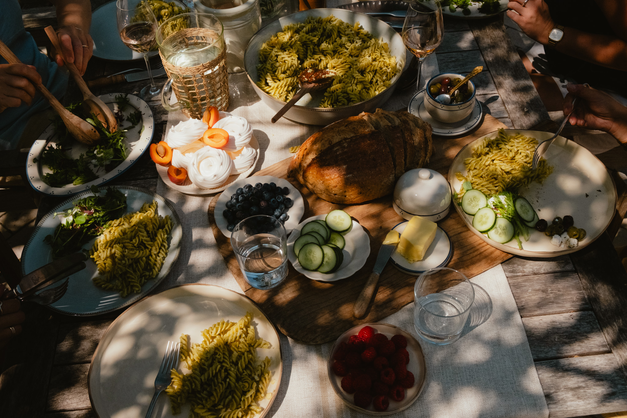An overhead view of a garden lunch spread — pesto pasta, sliced sourdough, meringues with apricots, blueberries and raspberries — with hands reaching in to serve.