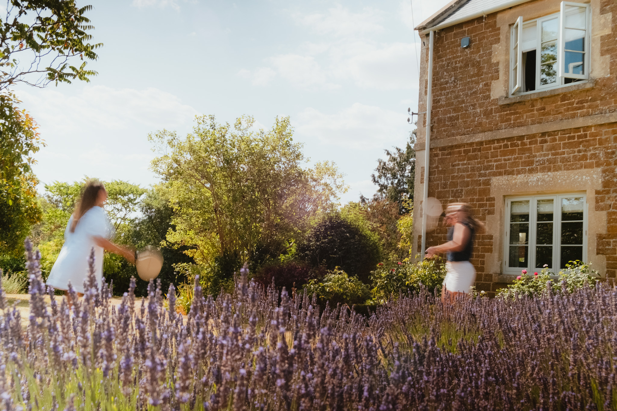 Two women in motion play garden games with wooden bats beside a honey-stone farmhouse, a lavender bed blurred in the foreground under bright summer light.