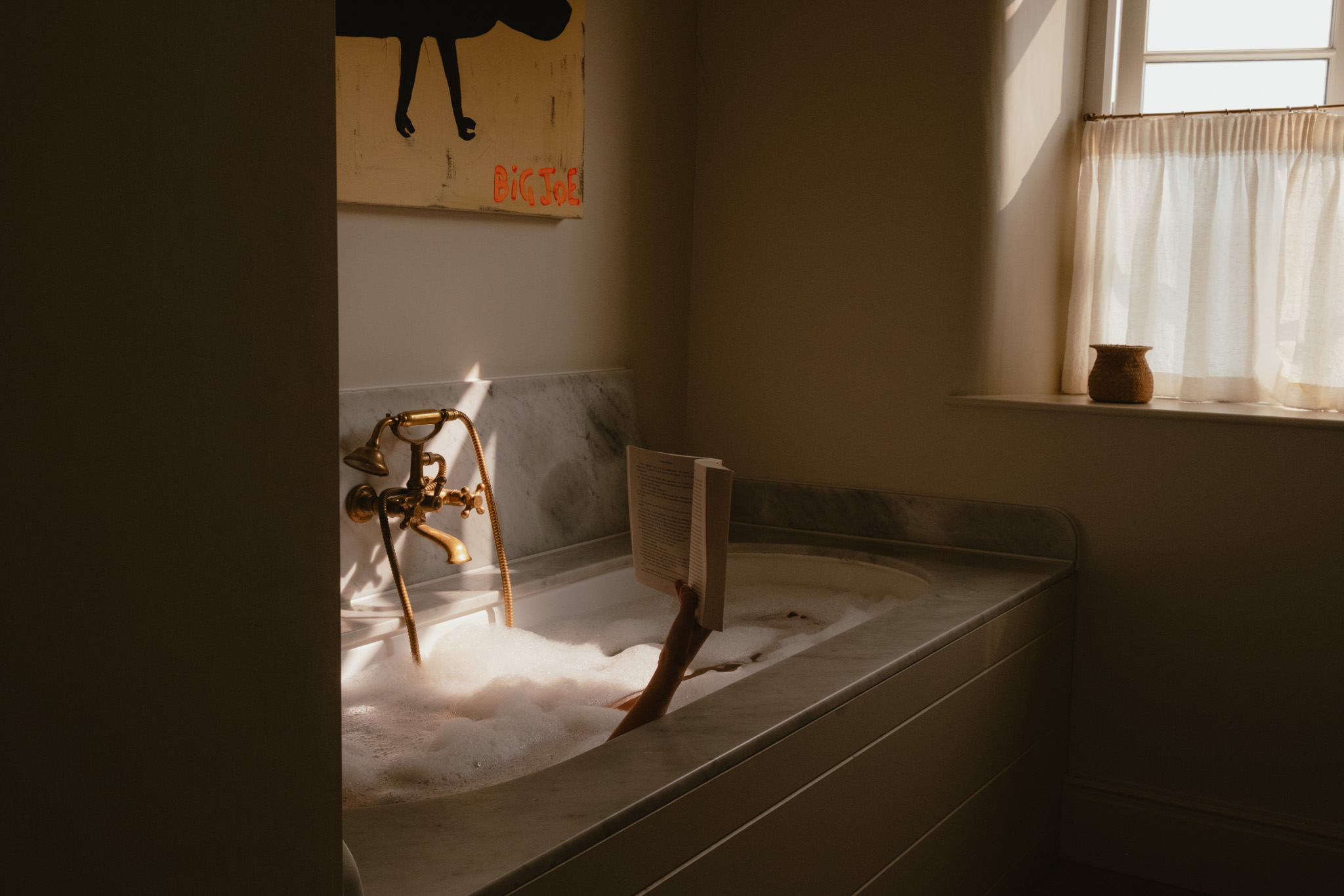A hand holding an open book rises from a bubble-filled bathtub, lit by a sunlit window with sheer curtains and a playful painting on the wall above brass taps.