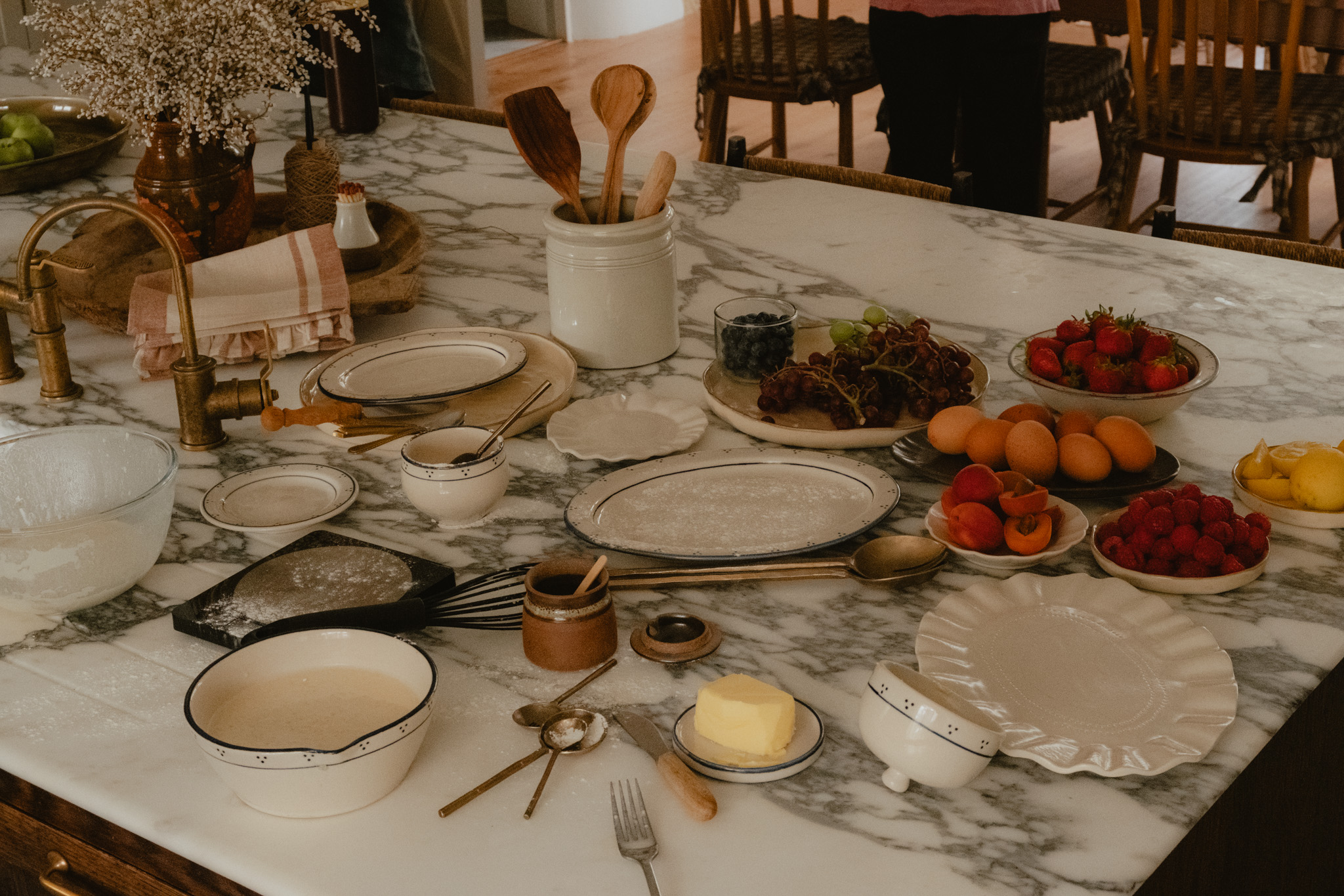 An overhead view of a marble kitchen island laid out for baking, with mixing bowls, wooden spoons, butter, eggs, berries and stone fruit gathered around a dusted pastry board.