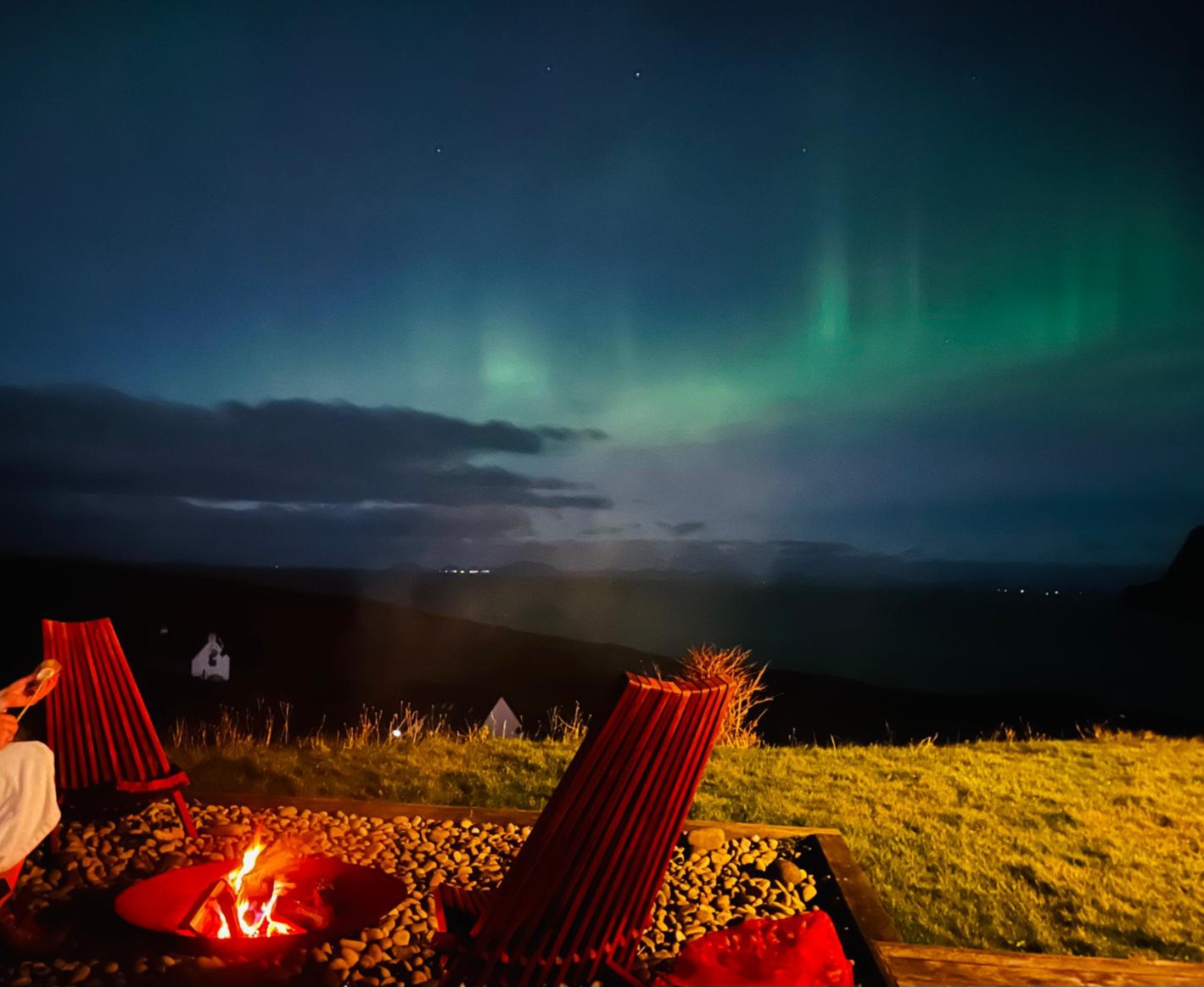 Aurora over the fire pit — Ribbons of green aurora rise above a glowing fire pit and empty red lounge chairs, the coastline and distant lights barely visible beneath the night sky.