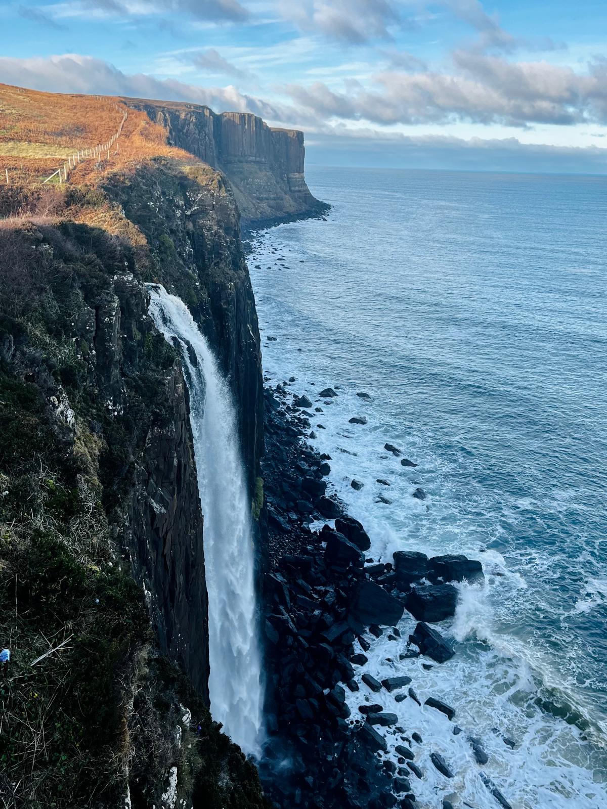 Kilt Rock waterfall — A slender waterfall drops in a single unbroken column down the dark basalt columns of Kilt Rock, meeting the crashing surf and scattered black boulders below a bright coastal sky.