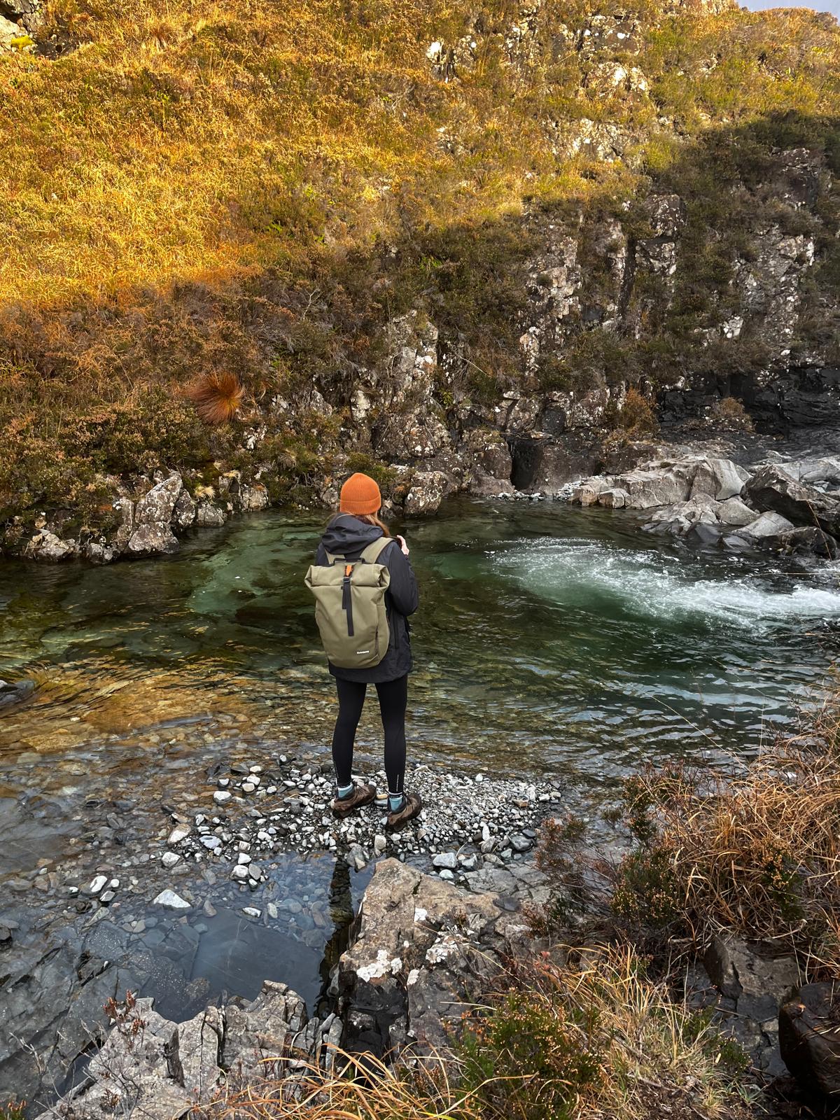 Hiker beside a Skye river pool — A hiker in a beanie and khaki backpack pauses on the rocks at the edge of a clear river pool, its green-black water swirling past sun-lit autumn grass and weathered stone banks.