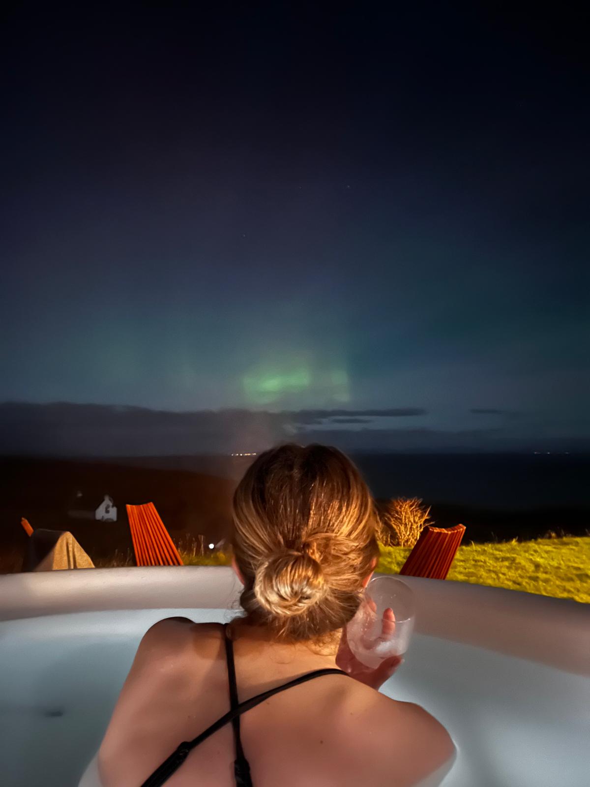 Hot tub under the northern lights — Seated in an outdoor hot tub with a drink in hand, a guest looks out over dark hills toward a faint green aurora glowing above the sea.