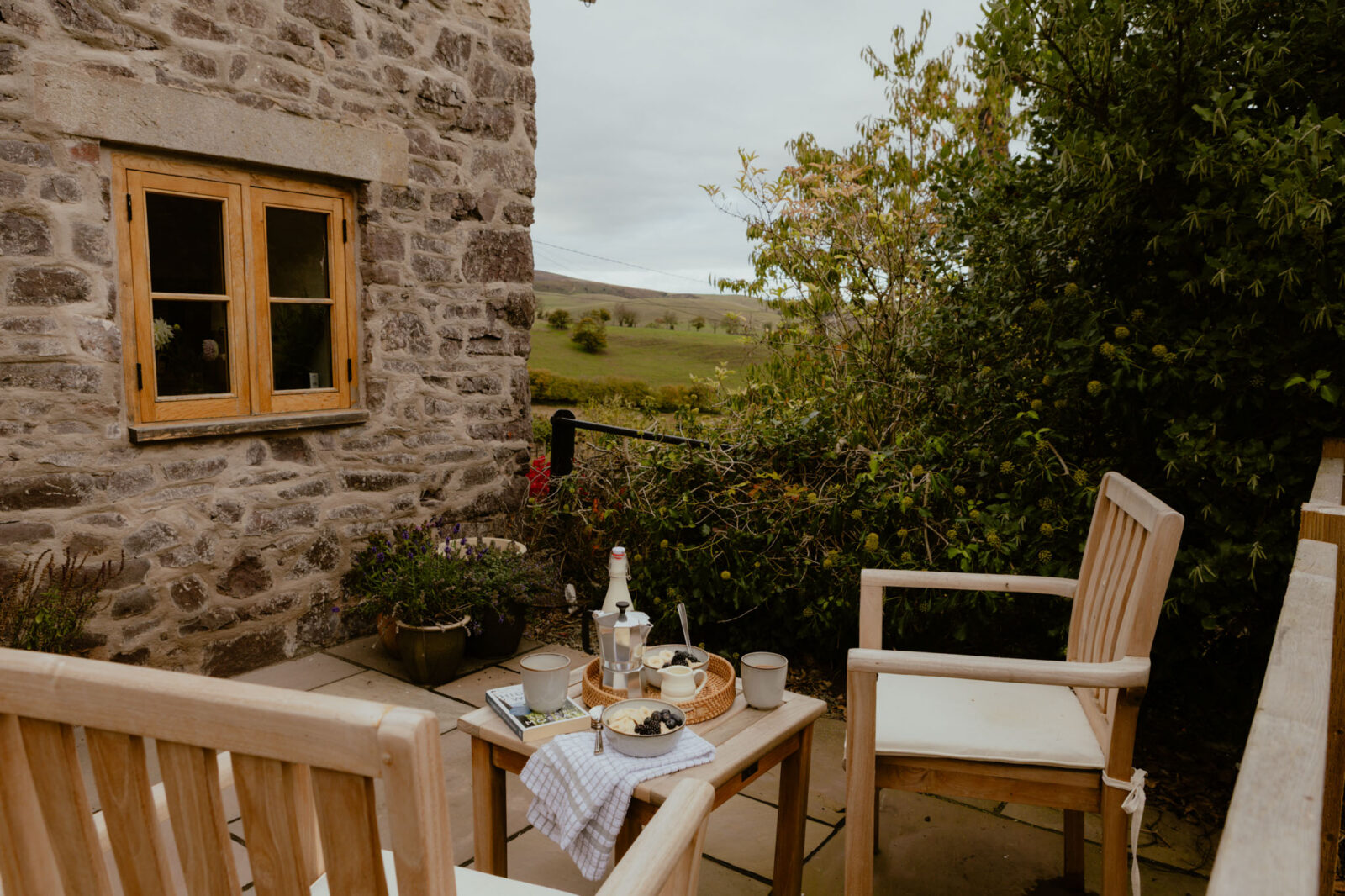 Breakfast for two on a stone patio beside the cottage wall: coffee mugs, a moka pot, and bowls of yoghurt with fruit, looking out over green hills.