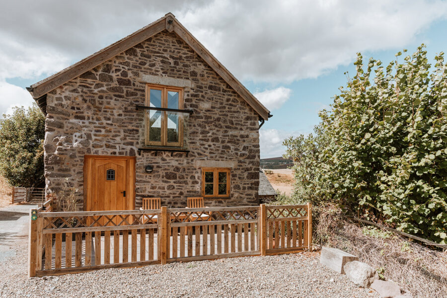 Exterior of a two-storey stone barn conversion with an oak front door and windows, a small patio with two wooden chairs behind a picket fence, and moorland hills in the distance.
