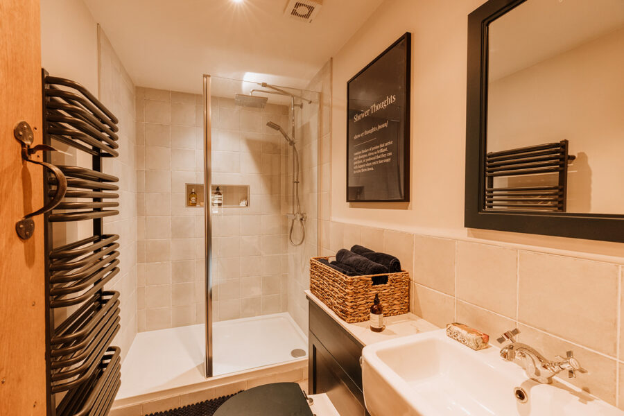 Compact shower room with a walk-in rainfall shower, beige tiles and a recessed niche, a black heated towel rail, a woven basket of navy towels on a dark vanity, and a framed “Shower Thoughts” print.