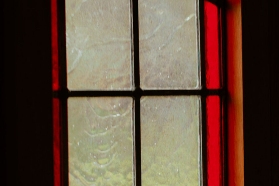 Small leaded window with red stained-glass edging glowing against a dark wall, its rain-speckled panes softly showing greenery outside.