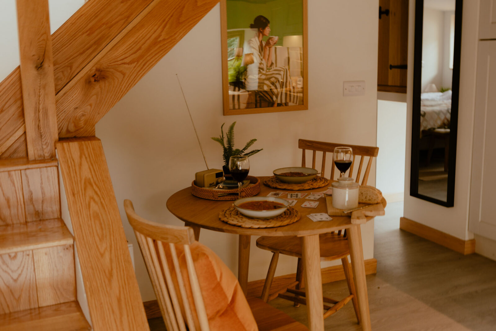 Cosy dining nook beside an oak staircase, with a round table set for two, a framed print on the wall, and a mirror reflecting the bedroom beyond.