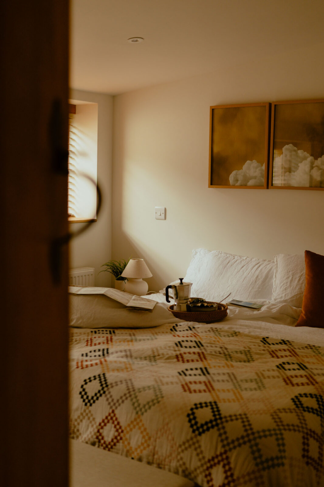 Glimpse through a half-open door into a bedroom bathed in soft light, with a patterned quilt, breakfast tray and coffee pot on the bed, and cloud artwork above.