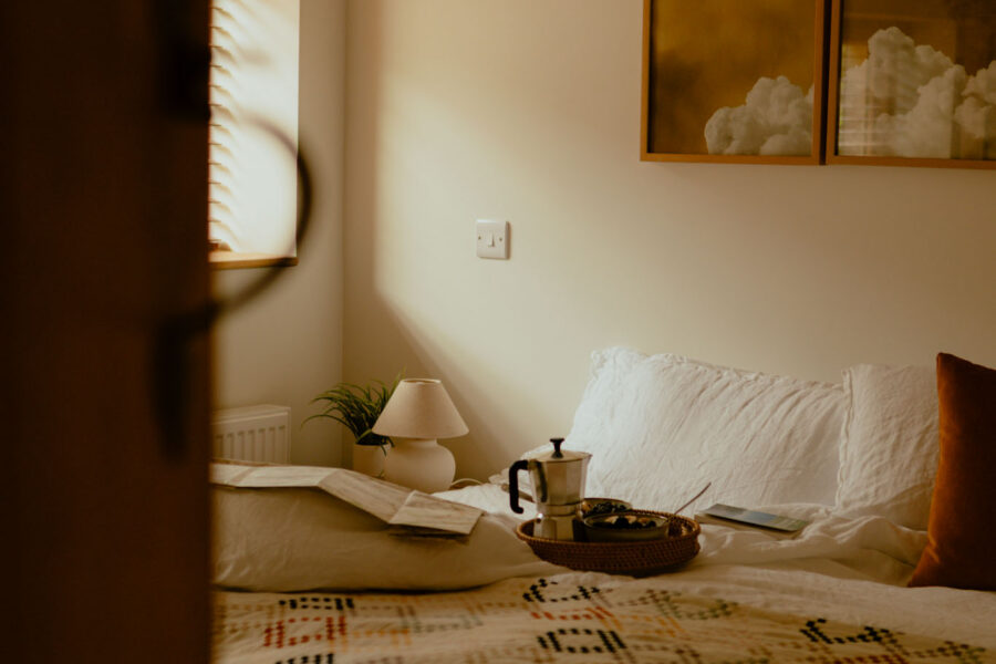 Glimpse through a half-open door into a bedroom bathed in soft light, with a patterned quilt, breakfast tray and coffee pot on the bed, and cloud artwork above.