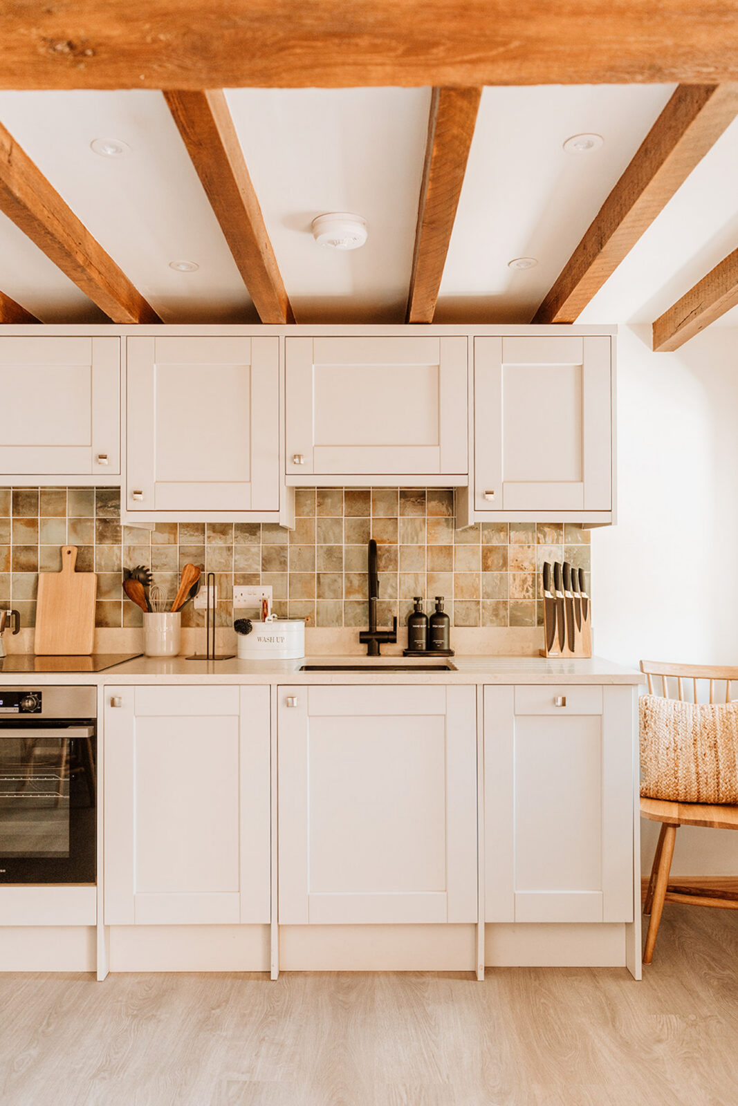 Cream shaker-style kitchen beneath exposed oak beams, with a green and amber tiled splashback, matte black tap, built-in oven, wooden utensils and a knife block, and a spindle-back chair to one side.