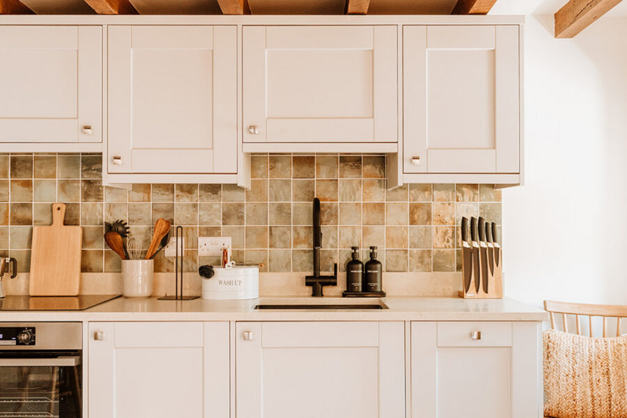 Cream shaker-style kitchen beneath exposed oak beams, with a green and amber tiled splashback, matte black tap, built-in oven, wooden utensils and a knife block, and a spindle-back chair to one side.