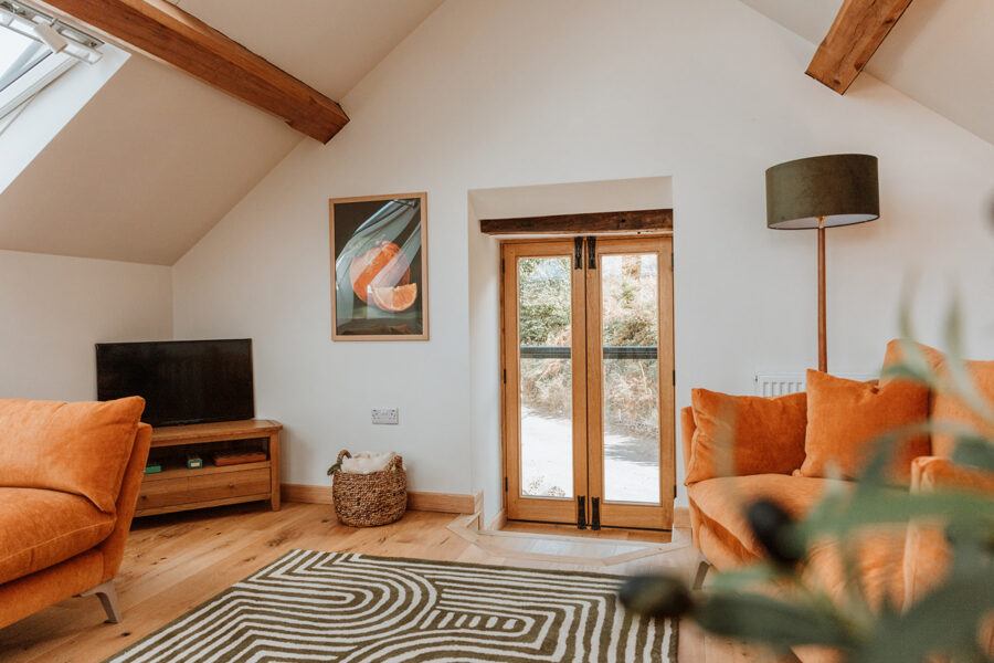 Bright vaulted living room with exposed oak beams, a skylight, two burnt-orange velvet sofas, a green and cream striped rug, a TV on an oak unit, a floor lamp, and glazed oak doors with a Juliet balcony.